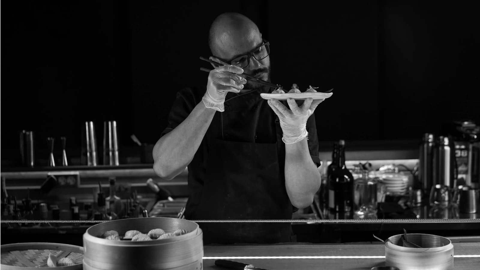 A chef plating dim sum with chopsticks at a bar counter, focused entirely on the dish