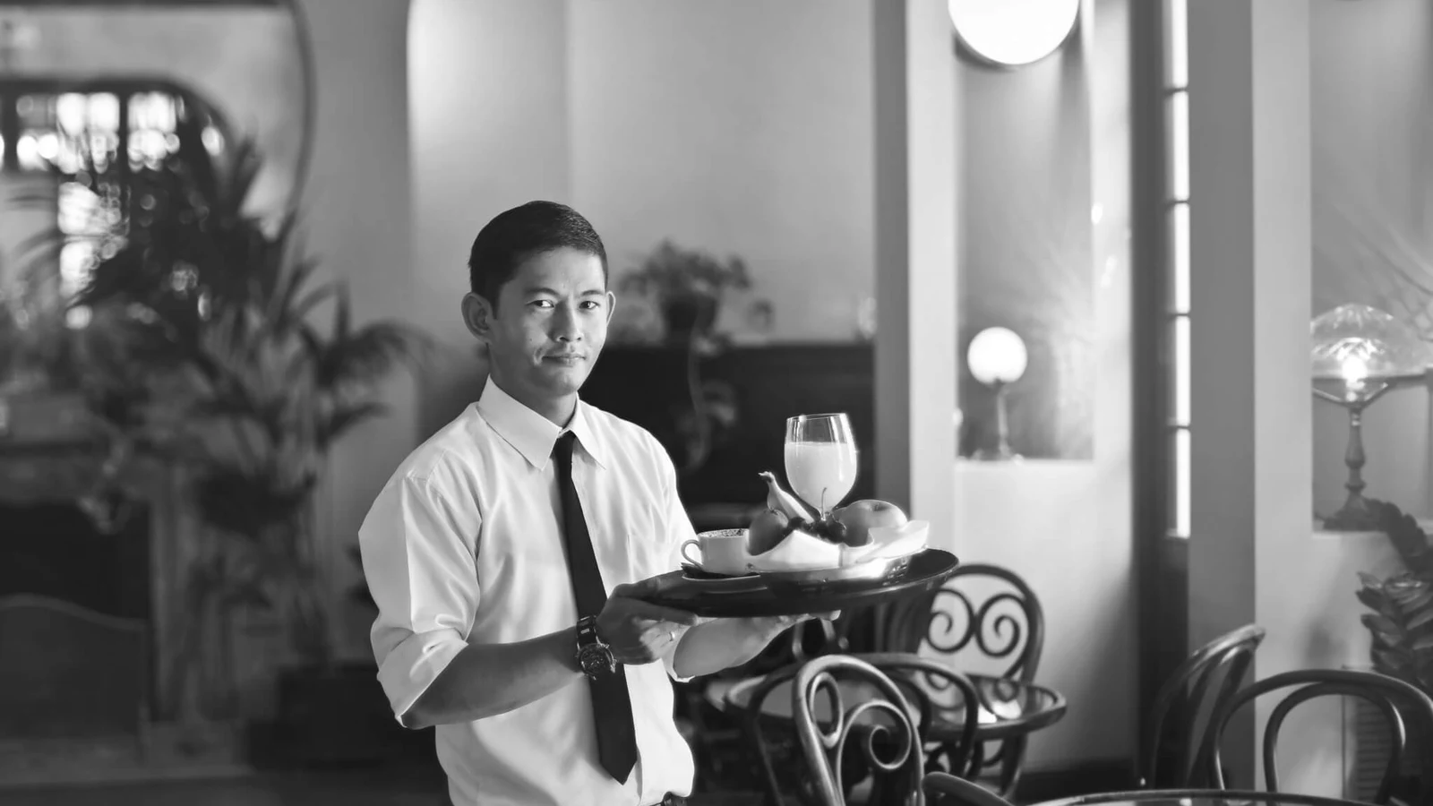 A waiter in a shirt and tie carrying a tray of drinks and fruit across a sunlit dining room